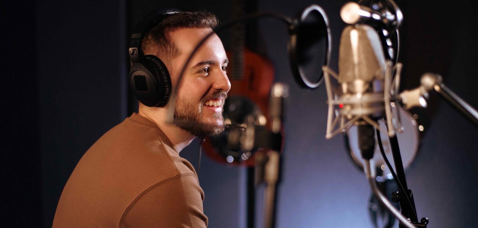 Homem jovem com barba curta e cabelo castanho usa fones de ouvido enquanto sorri em um estúdio de gravação. Ele veste uma camiseta marrom e segura um celular, sentado diante de um microfone profissional com pop filter. Ao fundo, instrumentos musicais desfocados compõem o ambiente. O homem é Rafael Witt, que se apresenta em São Paulo no próximo domingo 25 de maio.