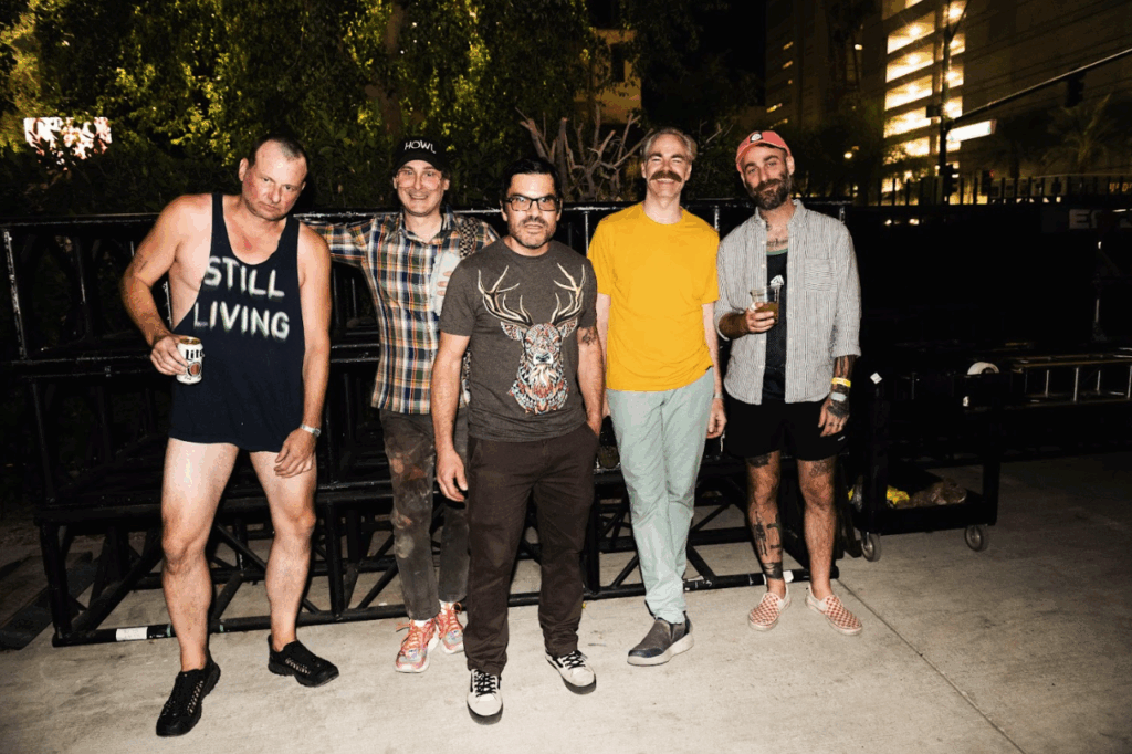 Integrantes da banda Cap’n Jazz nos bastidores de show noturno, com roupas casuais e bebidas em mãos