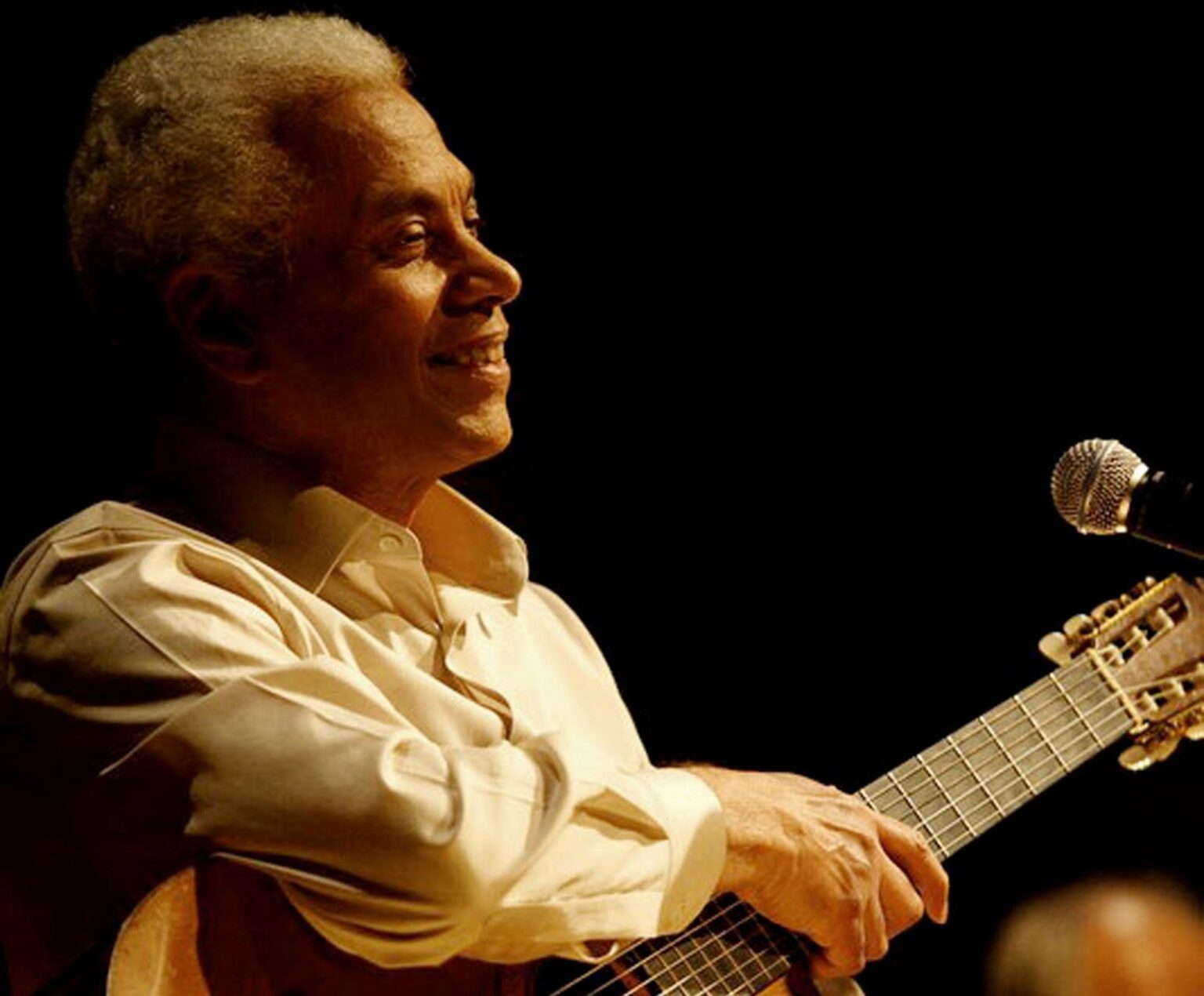 Paulinho da Viola sorri durante apresentação, segurando um violão e posicionado ao lado de um microfone, com iluminação suave em fundo escuro. Paulinho participará do Festival de Inverno 2025, com apresentação dia 2 de agosto.