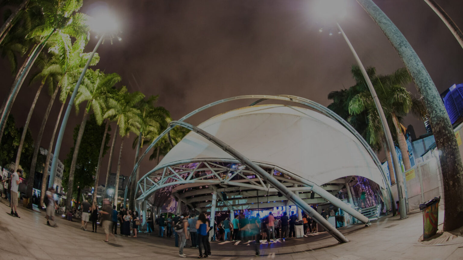 Foto noturna do Circo Voador, no Rio de Janeiro. A famosa tenda branca é sustentada por uma estrutura metálica curva, iluminada por luzes externas. Pessoas estão reunidas na entrada, sob palmeiras altas, criando uma atmosfera vibrante e acolhedora.