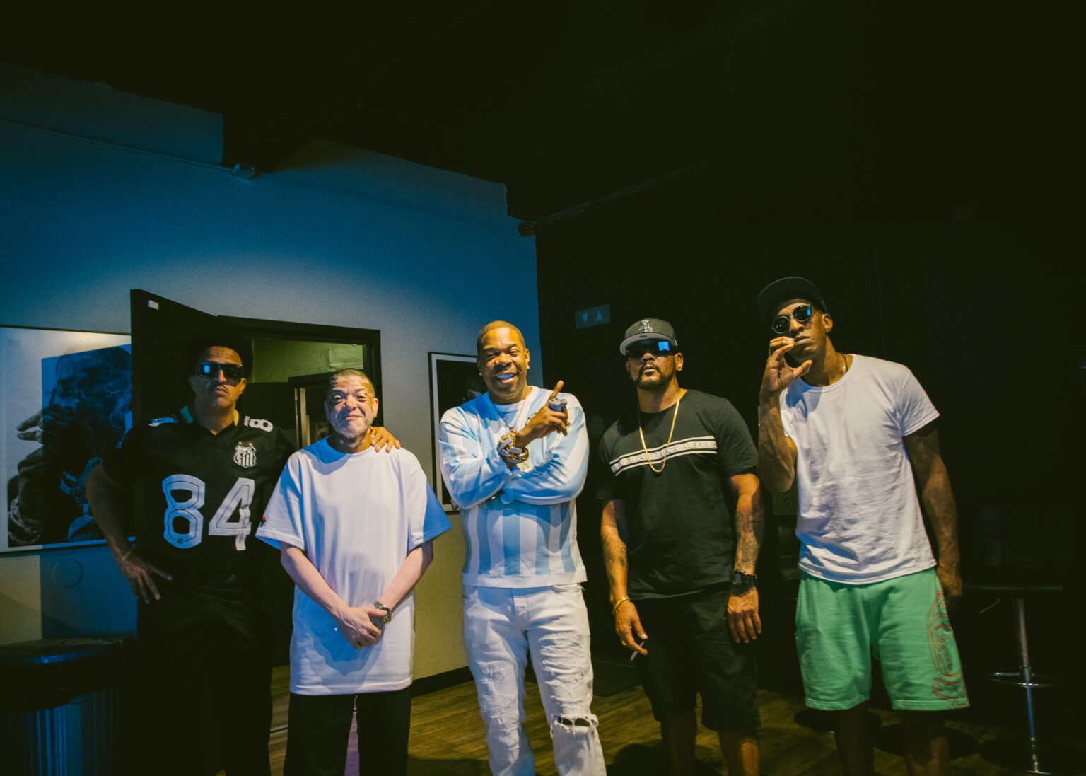Racionais MC’s e Busta Rhymes posam juntos em um estúdio com iluminação baixa. Todos usam roupas casuais e acessórios marcantes, como correntes e óculos escuros. O ambiente tem parede azul, quadros e uma porta aberta ao fundo. Foto: Jef Delgado.