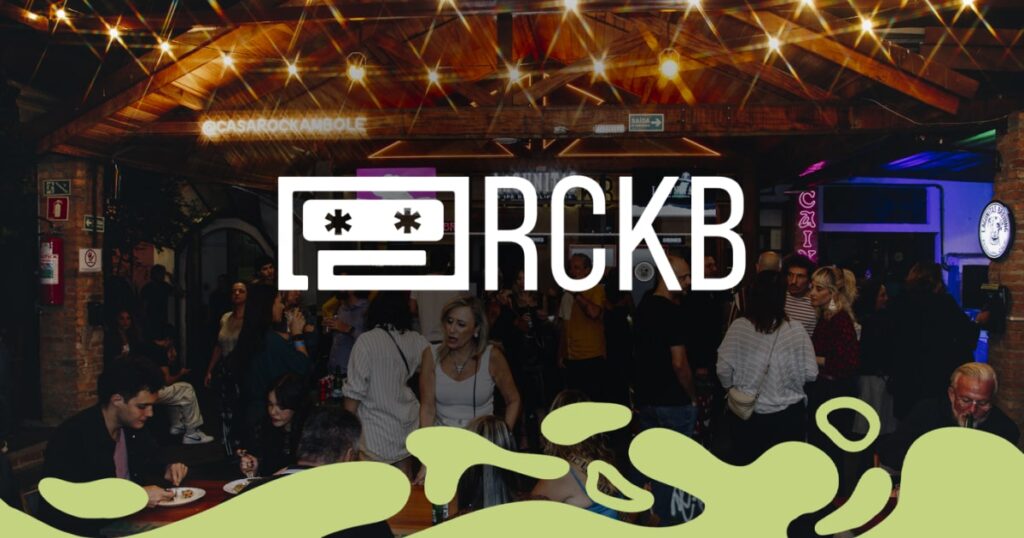 Fachada da Casa Rockambole (RCKB) com pessoas reunidas em ambiente descontraído à noite, sob luzes pendentes e decoração rústica.